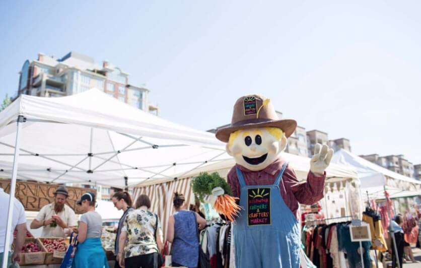A happy stuff scarecrow acting as a welcome guest to the farmers market which shows people selling their produce and hand made crafts. 