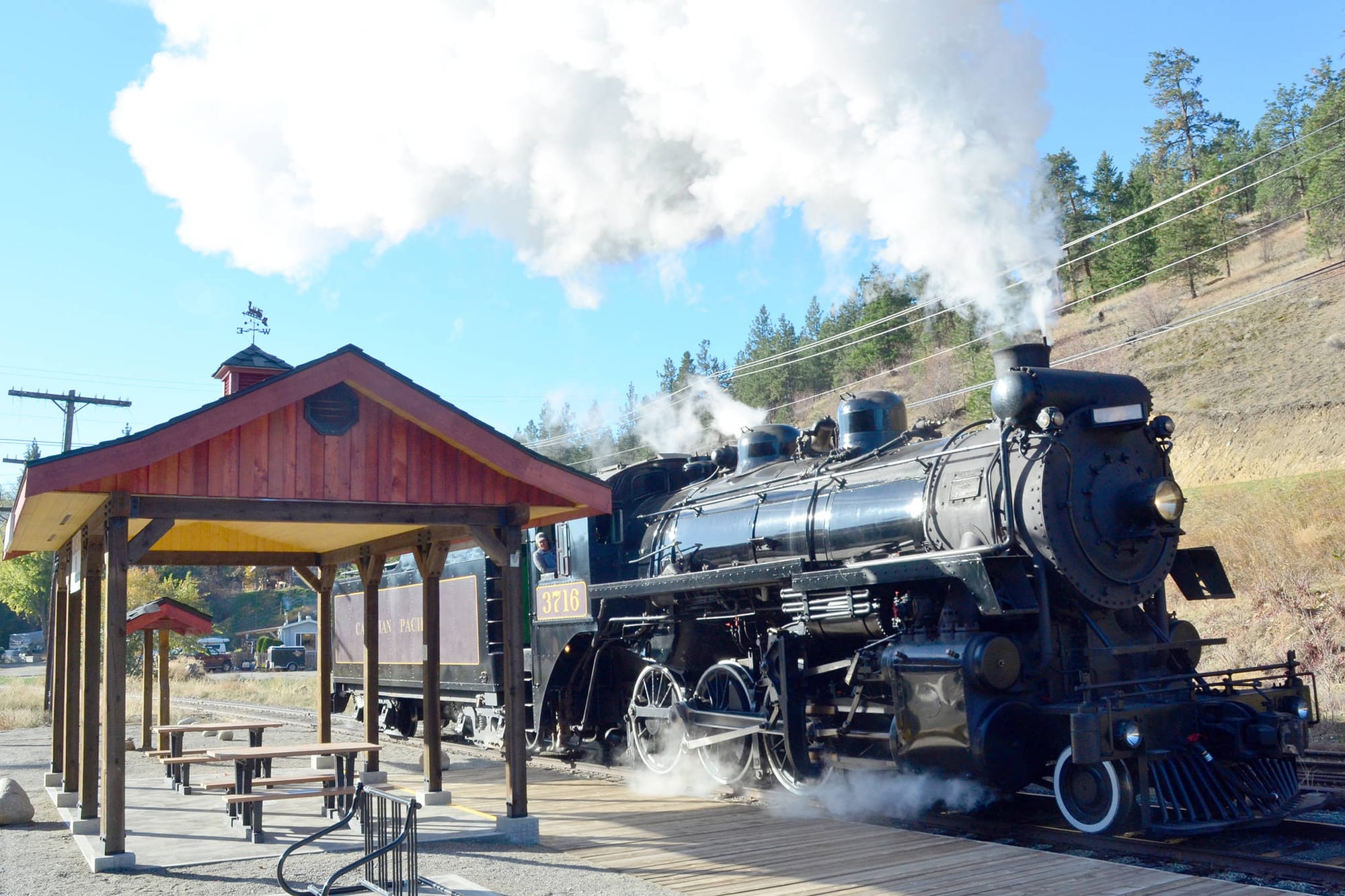 An old but well maintained and working steam train with steam coming from it's stacks and underneath, and is parked next to an old train wait station as you would have seen in the 1800's. 
