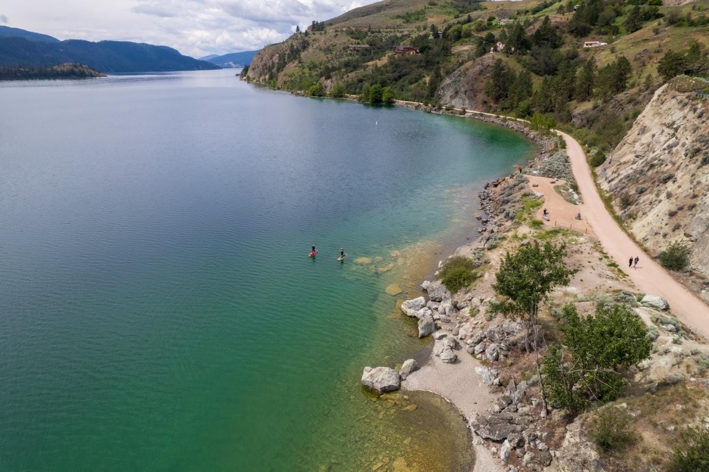Two people swimming in clear greenish water on the side of a rocky cliff and a hiking trail. 