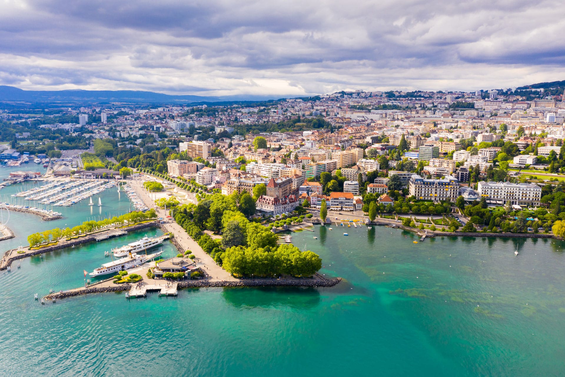  Aerial view of Lausanne, Switzerland, showcasing the city, marina, and turquoise waters of Lake Geneva under a cloudy sky.