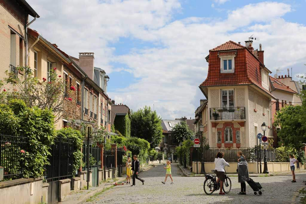 Charming residential street in the 20th arrondissement of Paris, France, with cobblestone pavement, greenery-lined houses, and people walking and cycling on a sunny day.