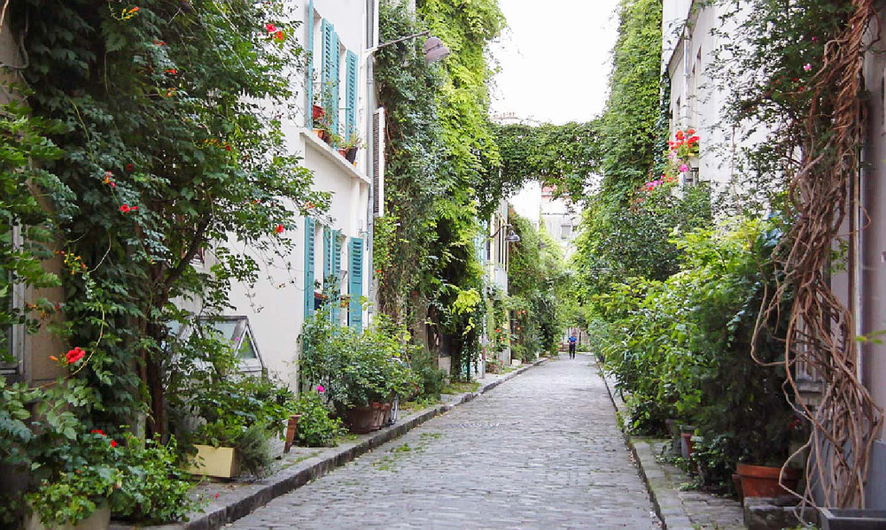 Charming cobblestone alley in Paris lined with ivy-covered buildings, colorful shutters, and lush greenery, creating a peaceful, garden-like atmosphere in the city.