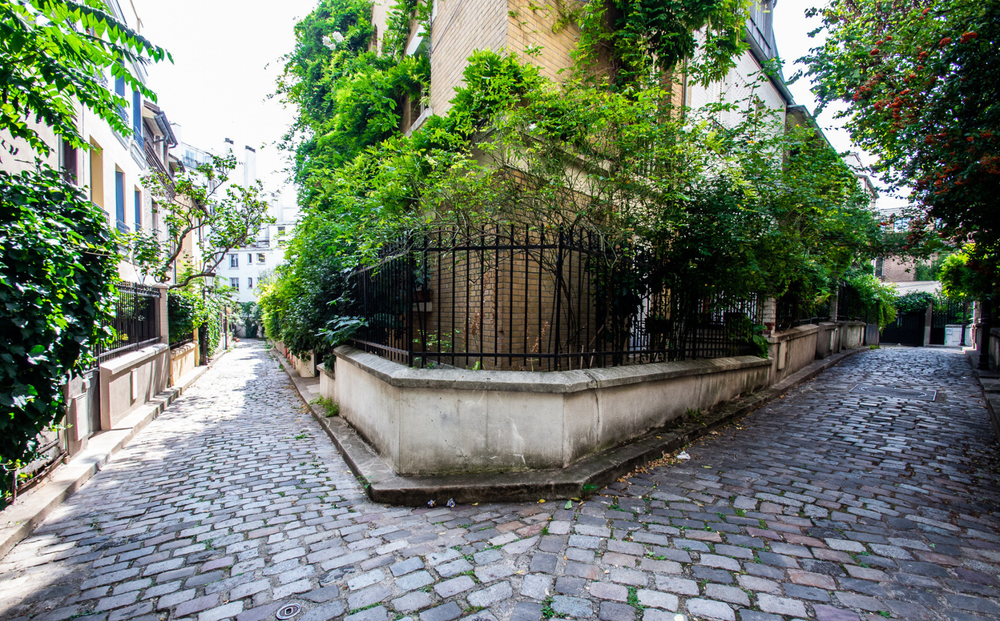 Peaceful cobblestone alley in Paris with intersecting paths, surrounded by ivy-covered buildings, wrought iron fences, and lush greenery on a sunny day.
