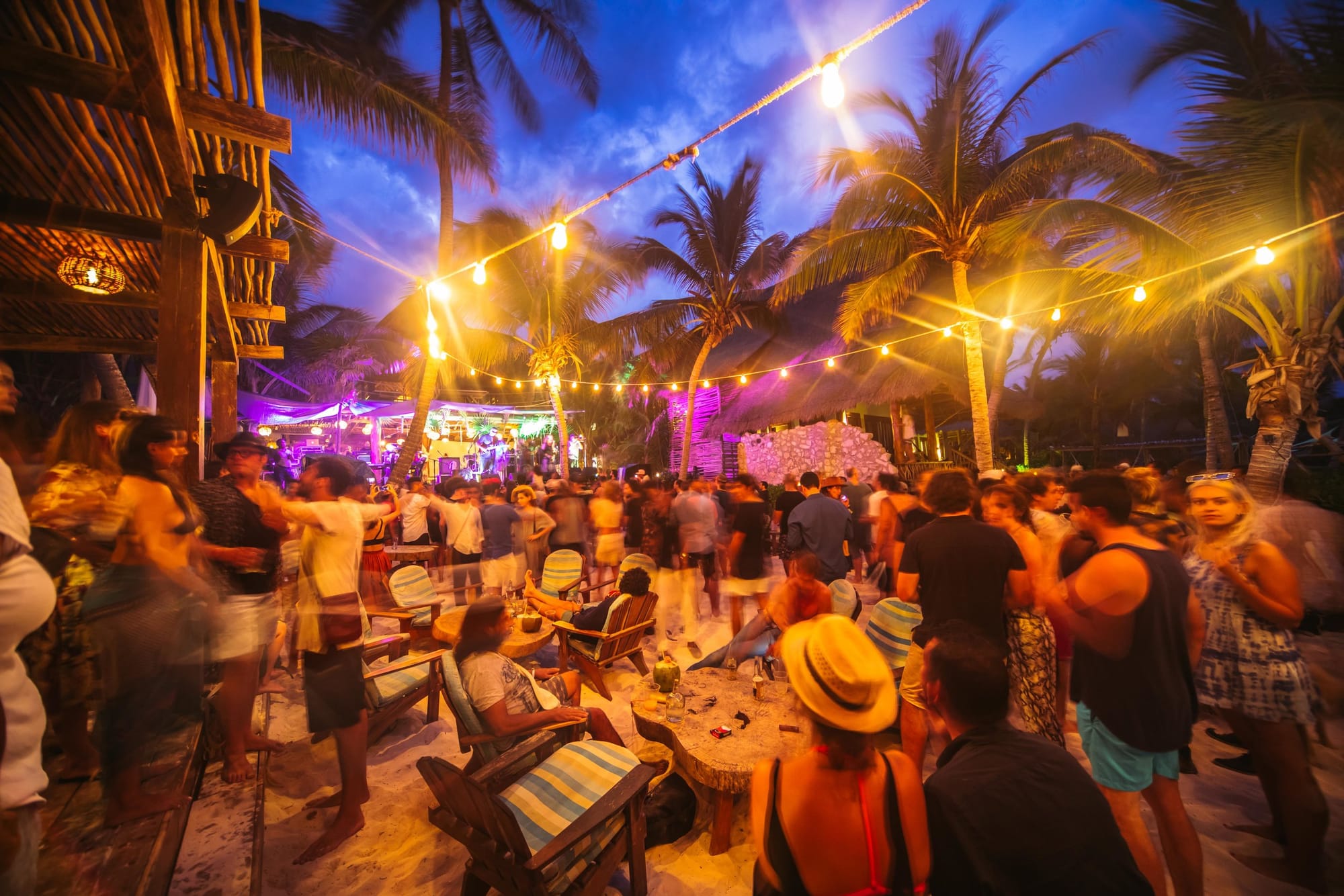 A lively beach party at the Art With Me Festival in Tulum, Mexico, with people dancing and socializing under warm string lights.