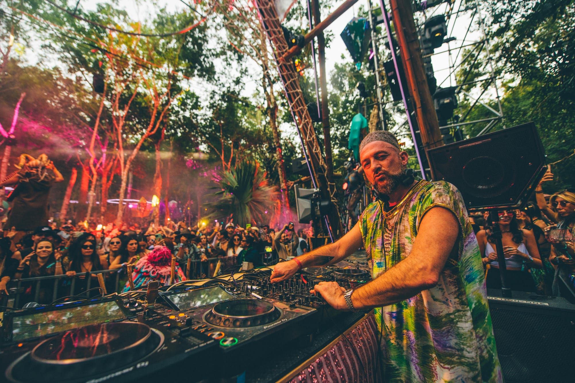 A DJ wearing a colorful, patterned outfit and a headscarf performs at Day Zero Festival in Tulum, Mexico, surrounded by a lush jungle setting.