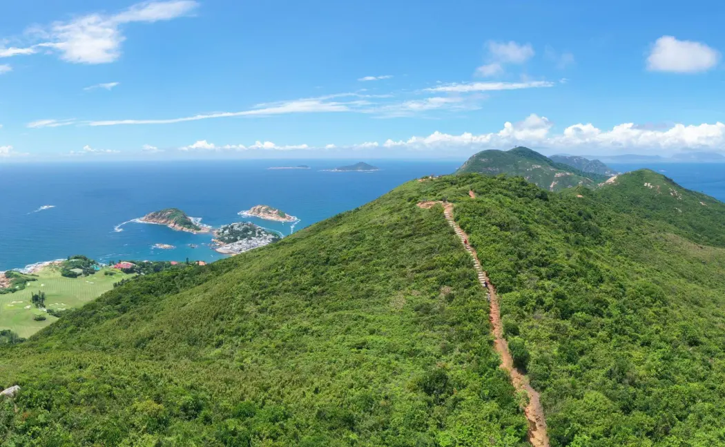 The Dragons Back trail in Hong Kong ariel view shows a mountain top covered in trees with a well worn trail running along the mountains peak.