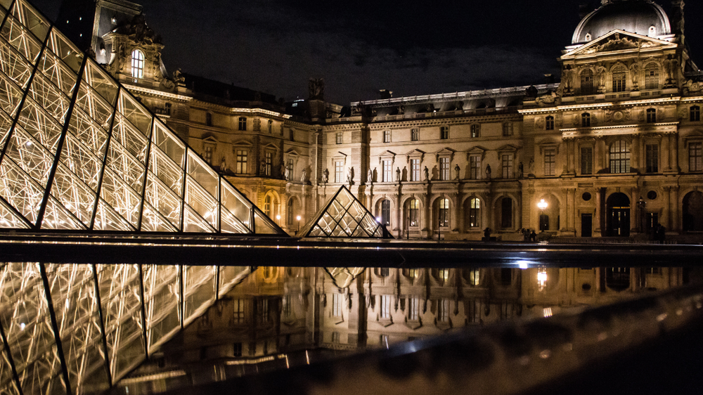 Illuminated glass pyramids and historic architecture of the Louvre Museum in Paris at night, with reflections in the water creating a symmetrical composition.
