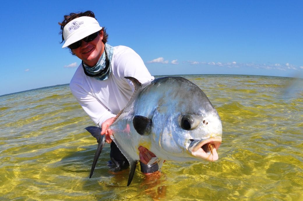 A man wearing a white long-sleeve shirt, sunglasses, and a visor holds a large permit fish in shallow, crystal-clear waters in Tulum, Mexico.