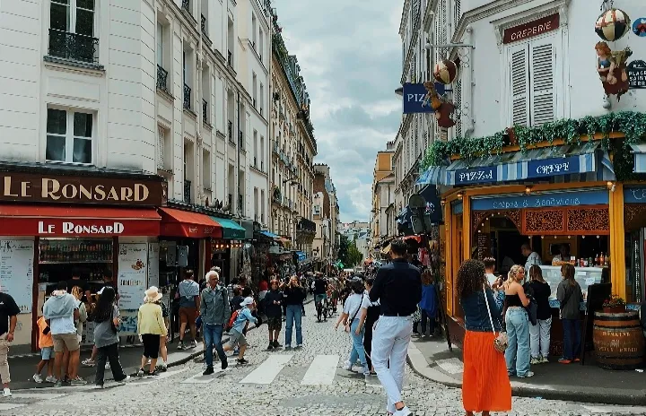 Bustling street scene in Montmartre, Paris, with people walking between cafés, restaurants, and crêperies on a cobblestone road lined with historic buildings.