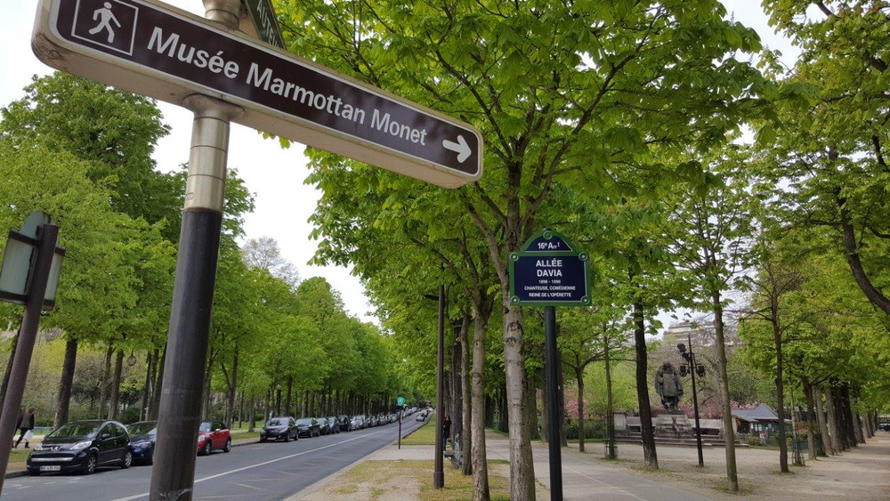 Street sign pointing toward Musée Marmottan Monet in Paris, France, along a tree-lined road with parked cars and pedestrians walking through Allée Davia in the 16th arrondissement.