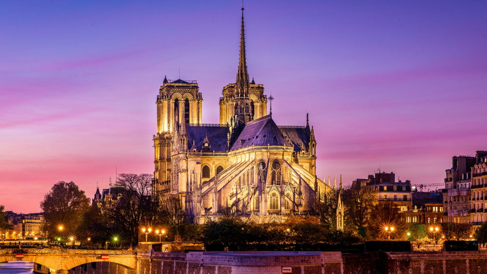 Notre-Dame Cathedral in Paris illuminated at sunset, with a vibrant purple and pink sky in the background and city lights reflecting along the Seine River.