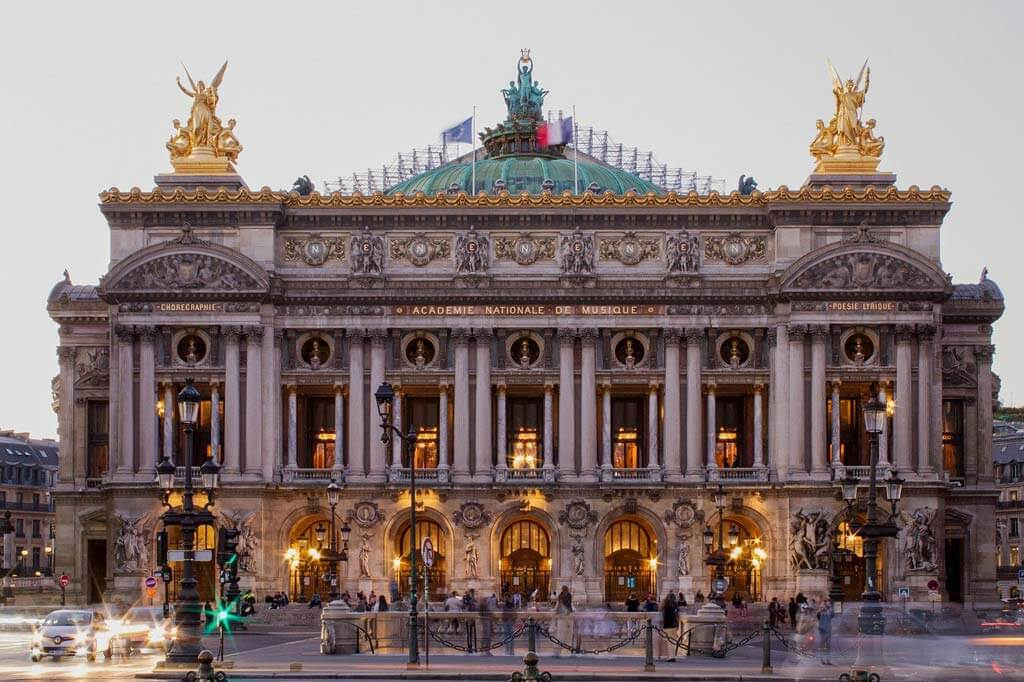 Front view of the Palais Garnier opera house in Paris, France, showcasing its ornate Beaux-Arts architecture, golden statues, and illuminated entrance at dusk.