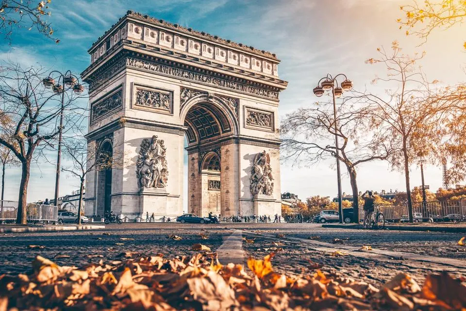 Arc de Triomphe in Paris, France, captured during autumn with fallen leaves in the foreground, golden sunlight, and bare trees lining the avenue.