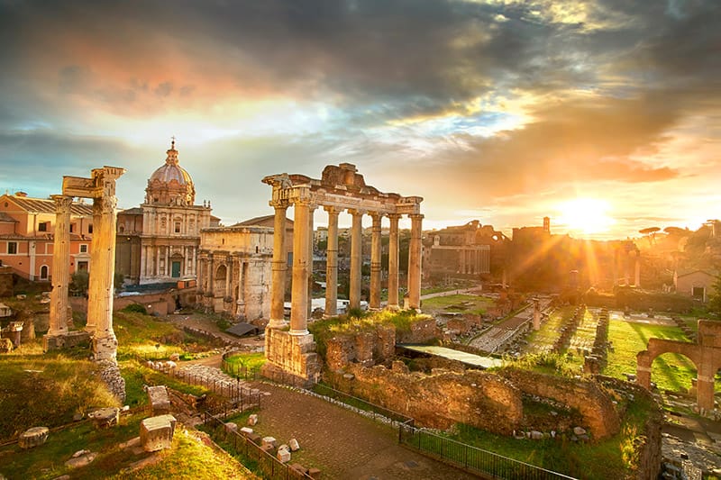 The ruins of the Roman Forum in Rome, Italy, bathed in the golden light of a setting sun. The image showcases towering ancient columns, remnants of historic structures, and the dome of a nearby church in the background. 