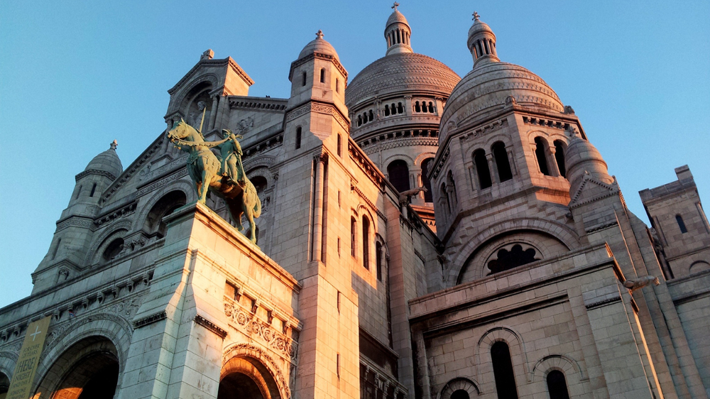 Close-up view of the Sacré-Cœur Basilica in Paris, France, with its domes and equestrian statue glowing in warm sunset light against a clear blue sky.
