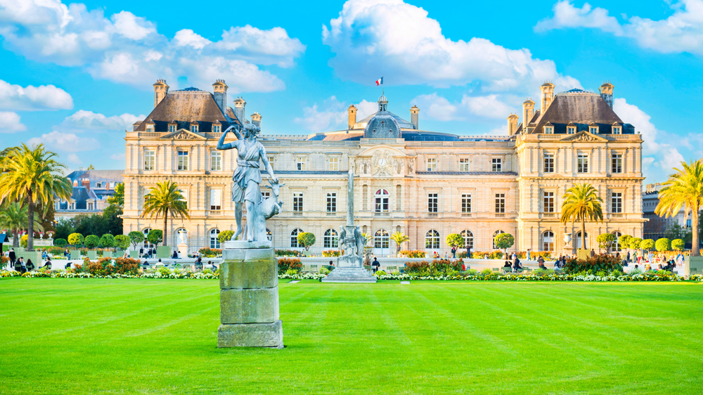 Statue and manicured lawn in front of the Luxembourg Palace in the Luxembourg Gardens, Paris, on a bright day with blue skies and scattered clouds.