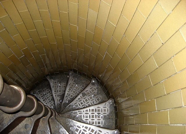  A narrow, winding spiral staircase inside St. Peter’s Basilica dome at the Vatican in Rome. The steep metal steps have a perforated design, enclosed by curved yellow-tiled walls. A central handrail provides support for climbers navigating the confined space as they ascend or descend within the historic structure.