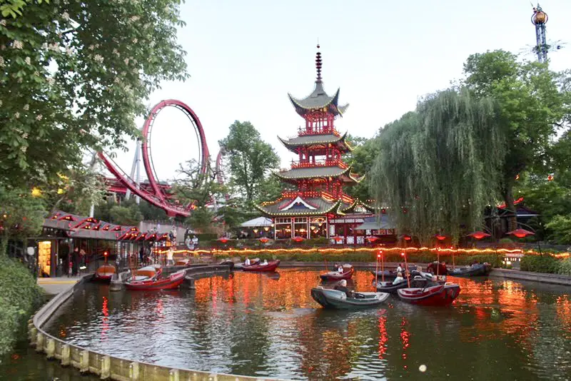 Boats floating in the water and a roller coasters in the background near what appears to be a Japanese style building at Tivoli Gardens Amusement park in Copenhagen, Denmark.
