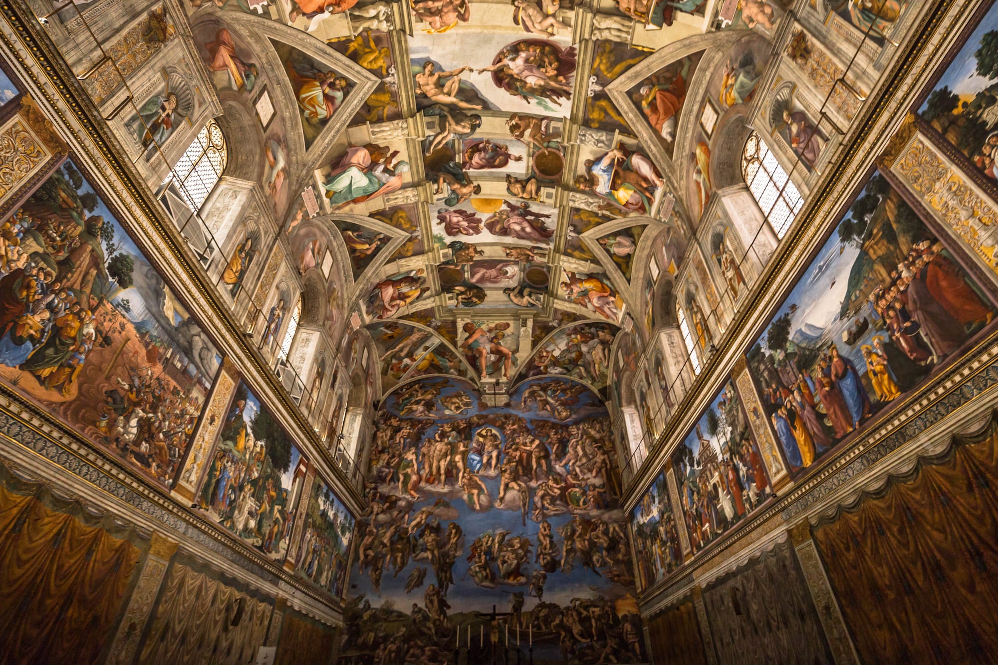 The interior of the Sistine Chapel in Vatican City, showcasing Michelangelo's famous frescoes on the ceiling and the Last Judgment on the altar wall. The ceiling features intricate biblical scenes, including The Creation of Adam, while the side walls display Renaissance-era frescoes depicting religious narratives. 