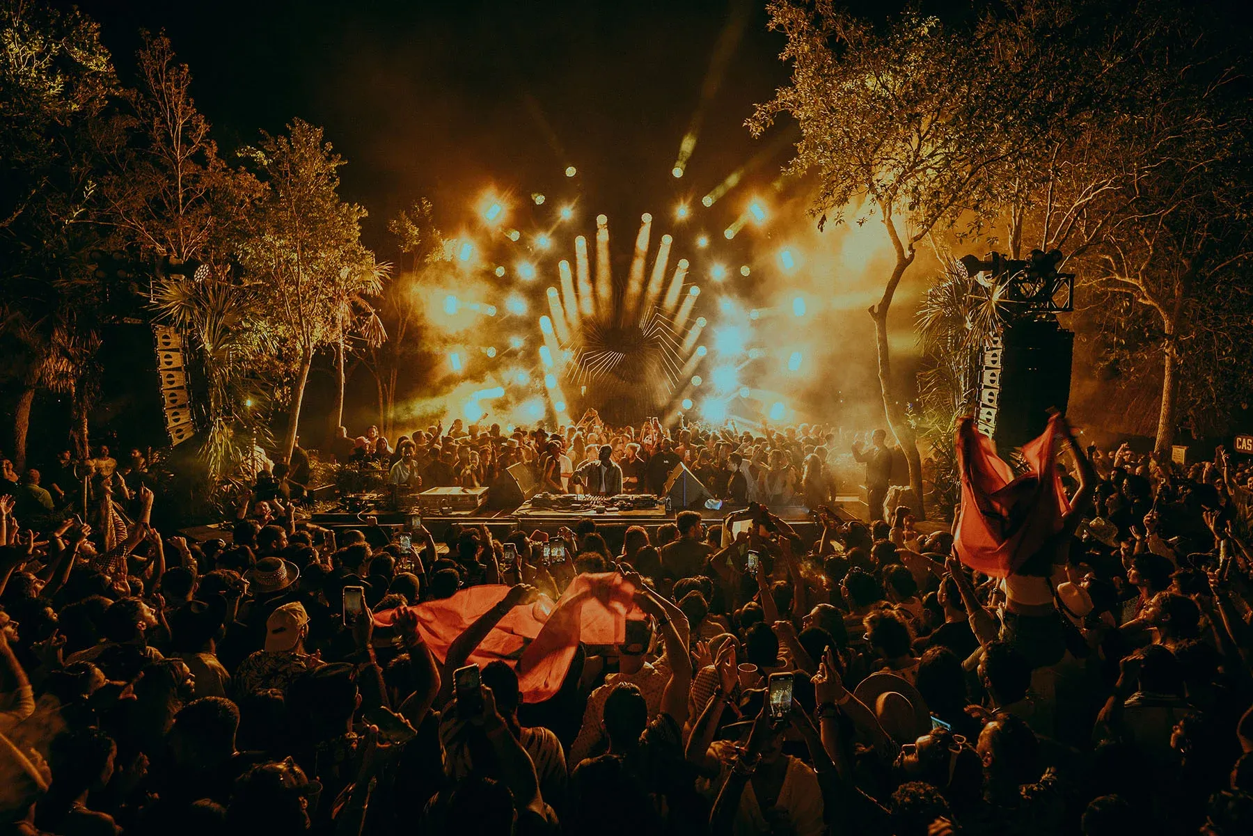 A vibrant nighttime scene at Zamna Festival in Tulum, Mexico, with a large crowd gathered around an outdoor stage. A DJ performs at the center, surrounded by an array of bright golden stage lights illuminating the tropical setting.