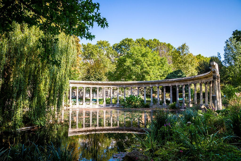 The classical colonnade and reflecting pond in Parc Monceau, Paris, surrounded by lush greenery and trees under a clear blue sky.