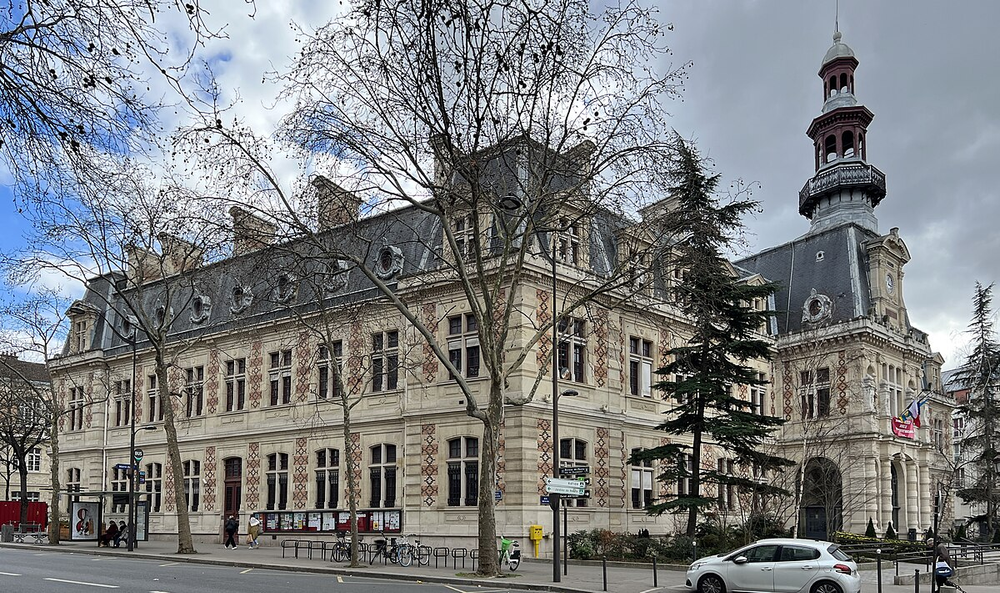 Exterior of the historic 14th arrondissement town hall (Mairie du 14e) in Paris, France, featuring ornate stonework, a tall bell tower, and a mix of cloudy and blue skies overhead.