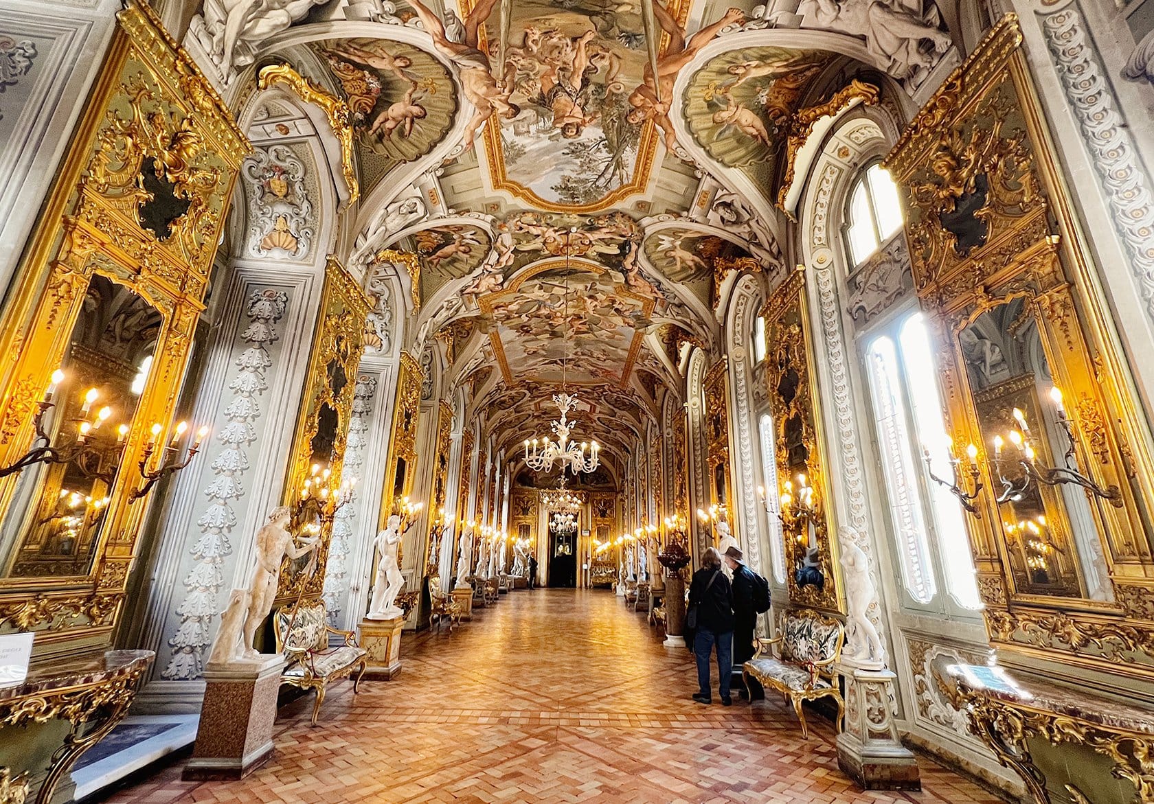 A lavishly decorated hallway inside the Doria Pamphilj Gallery in Rome, Italy, featuring an opulent Baroque-style ceiling adorned with intricate frescoes of mythological figures. The long, ornate corridor is lined with gilded mirrors, chandeliers, marble statues, and antique furniture, with soft candlelight adding to the grandeur.