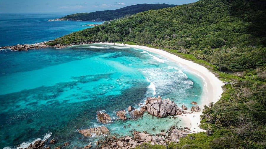 Aerial view of Anse Cocos Beach in La Digue, Seychelles, showcasing white sand, turquoise waters, granite rock formations, and lush tropical forest along the shoreline.