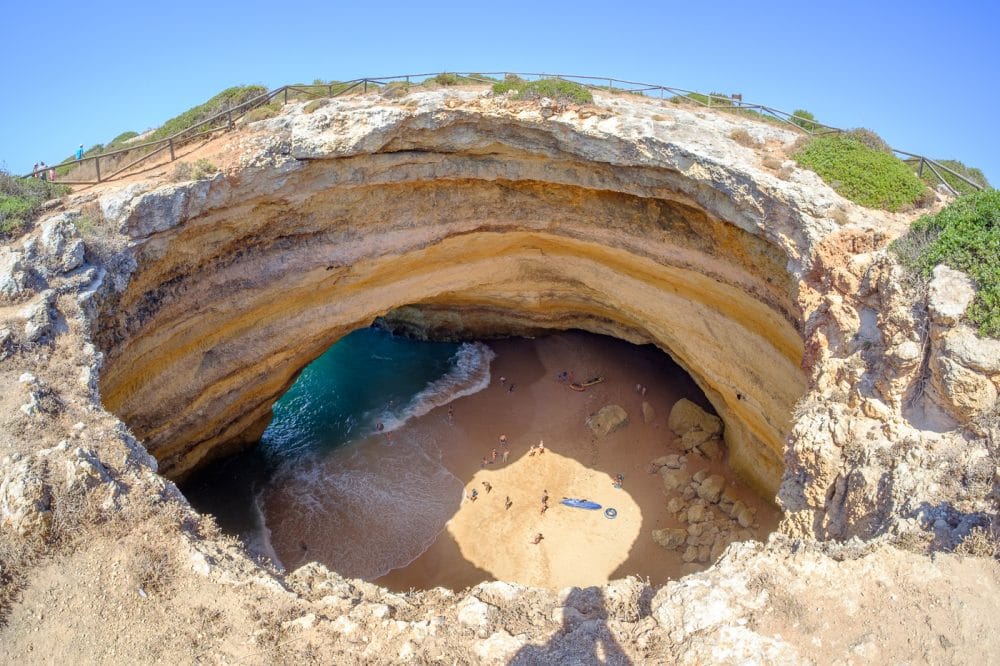 Aerial view of the Benagil Sea Cave in Algarve, Portugal, showing the natural rock dome with a circular skylight opening, golden sand beach, and turquoise water inside the cave.
