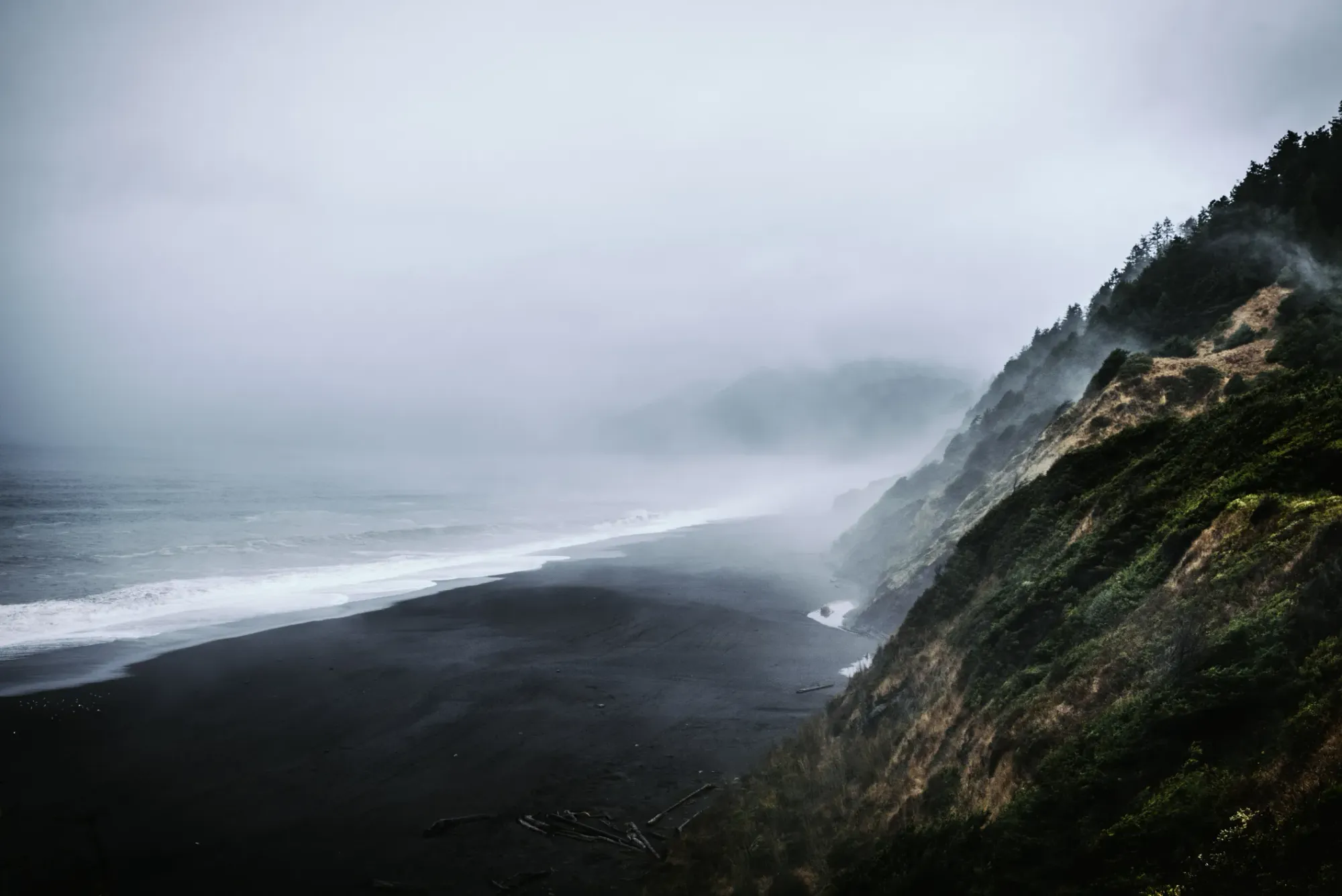 Misty coastal view of Black Sands Beach in California, featuring dark sand, ocean waves, and rugged cliffs covered in greenery under a moody, overcast sky.