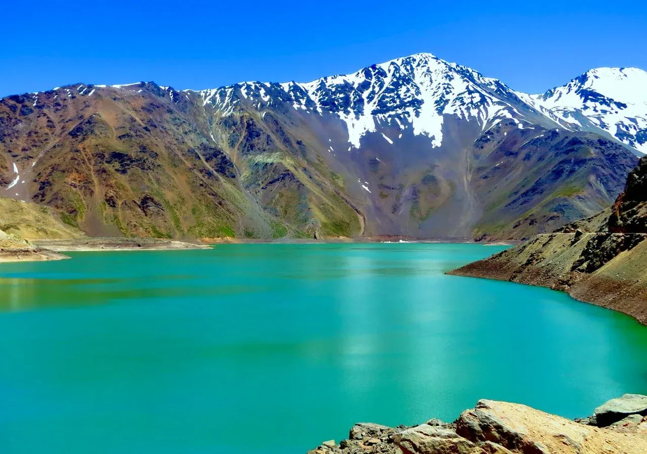 A large body of pretty water sitting down in a canyon at the base of a snow covered mountain peak.