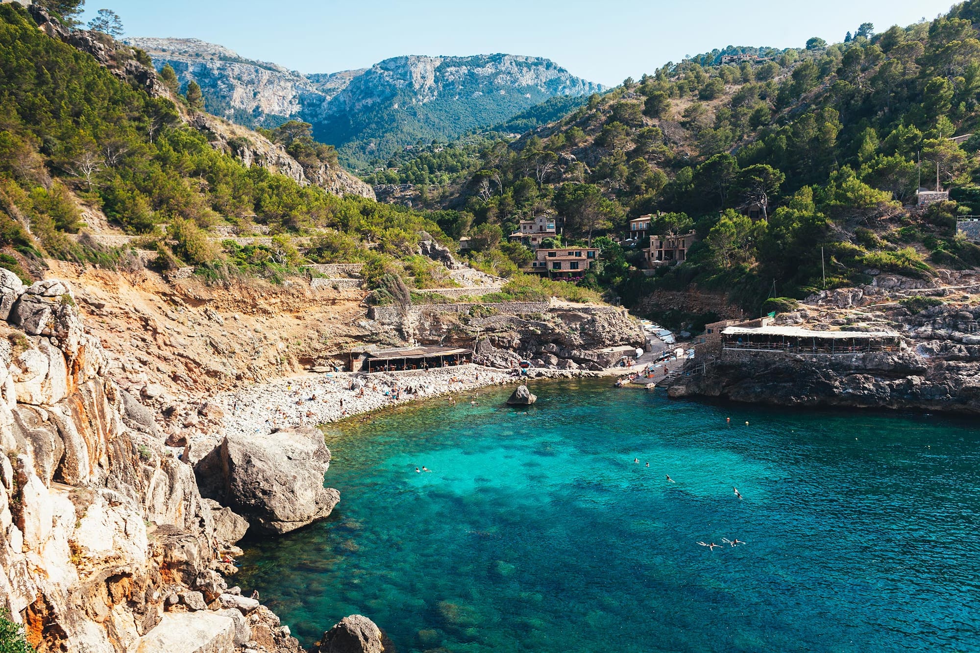 Secluded cove with turquoise water, surrounded by cliffs and greenery. People swim and relax on the pebbly beach, with small buildings on the right