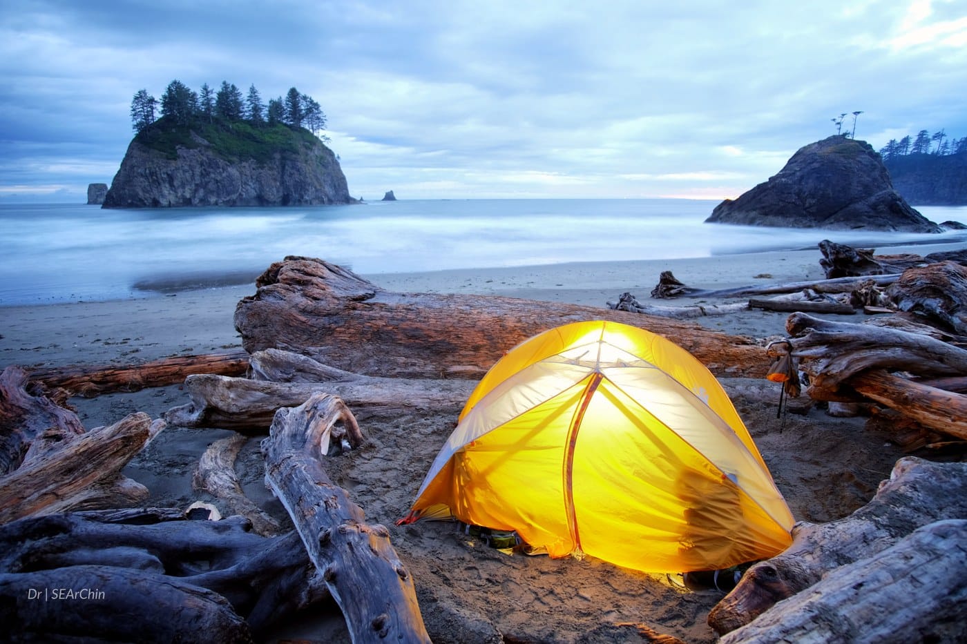 Glowing yellow tent set up on the driftwood-covered shore of Second Beach in Olympic National Park, Washington, with sea stacks and forested cliffs in the background under a moody sky.