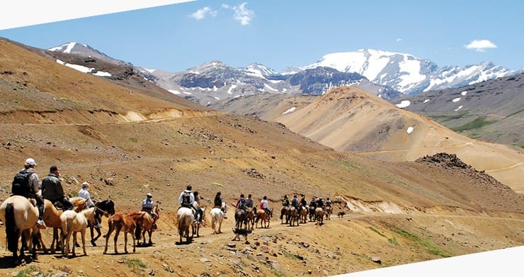 People riding horses in a strait line down a barren mountain size, and the mountains in the distance have snow covered peaks.