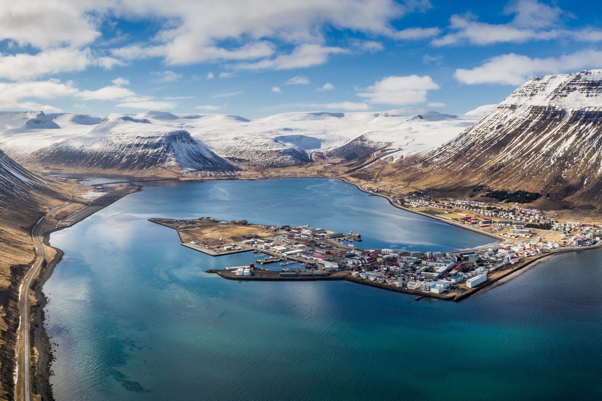 Aerial view of Ísafjörður, a coastal town in Iceland, nestled between snow-capped mountains and a calm fjord, with colorful buildings and a small harbor extending into the turquoise waters.