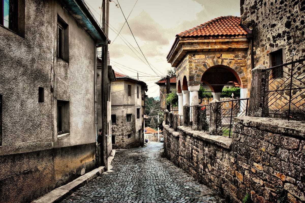 Narrow cobblestone street in Kratovo, North Macedonia, lined with old stone and plaster buildings, leading to a historic arched structure with red roof tiles and white columns, under a cloudy sky.