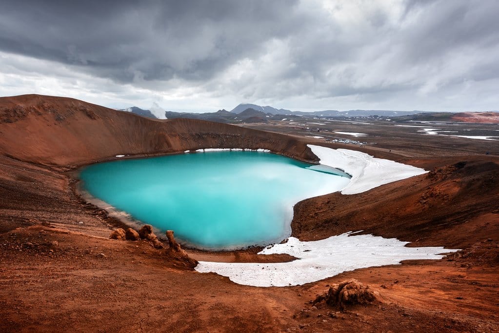 Volcanic crater lake with striking turquoise water surrounded by red-brown rocky terrain and patches of snow, under a dramatic cloudy sky near Lake Mývatn in northern Iceland.
