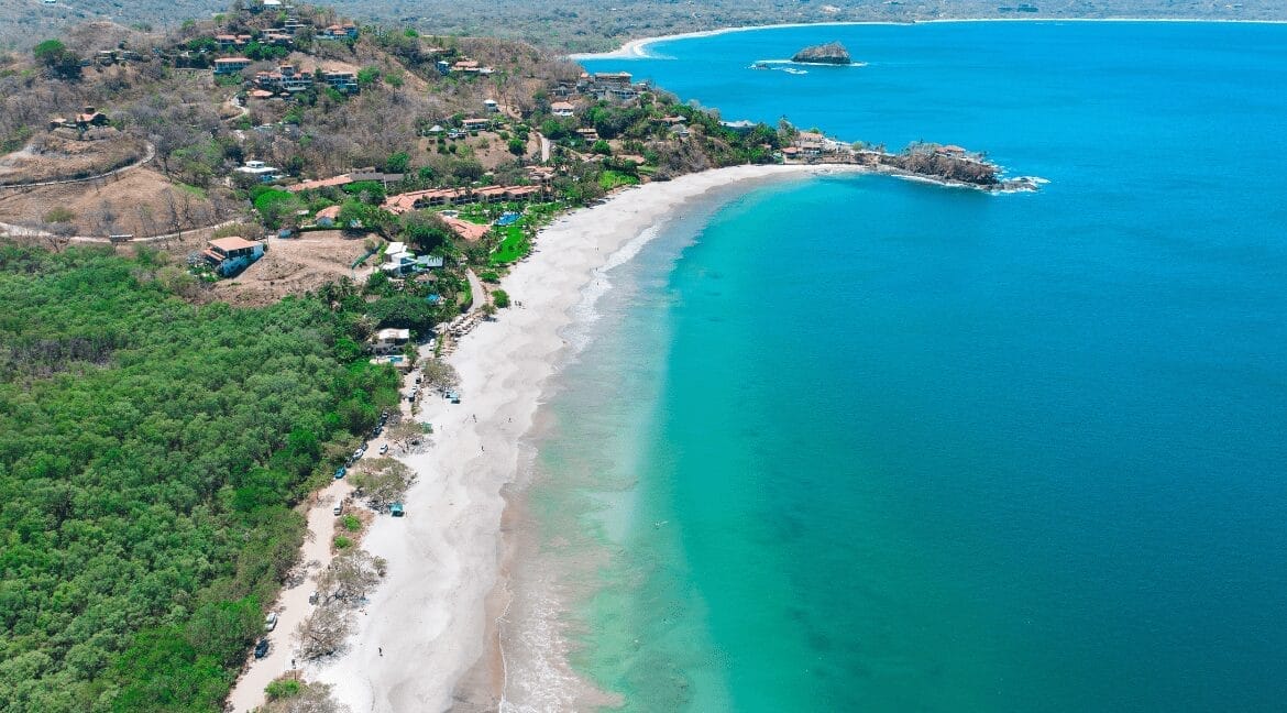 Aerial view of Playa Mina Beach in Costa Rica, featuring a crescent-shaped shoreline with soft white sand, turquoise waters, coastal greenery, and hillside villas overlooking the Pacific Ocean.