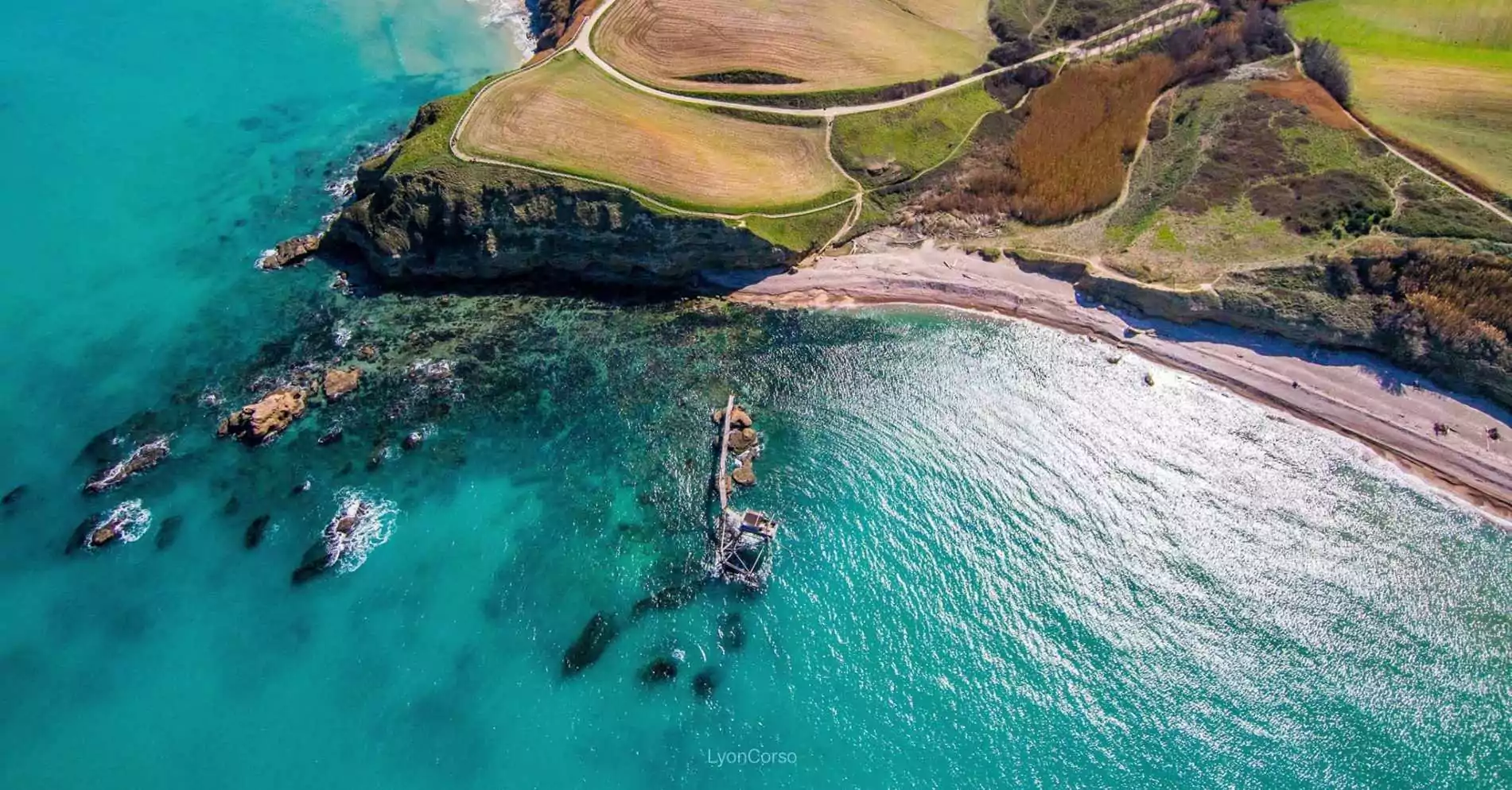 Aerial view of Punta Aderci Beach in Italy, showcasing turquoise waters, a natural coastline with cliffs and fields, and a traditional trabocco fishing platform extending into the Adriatic Sea.