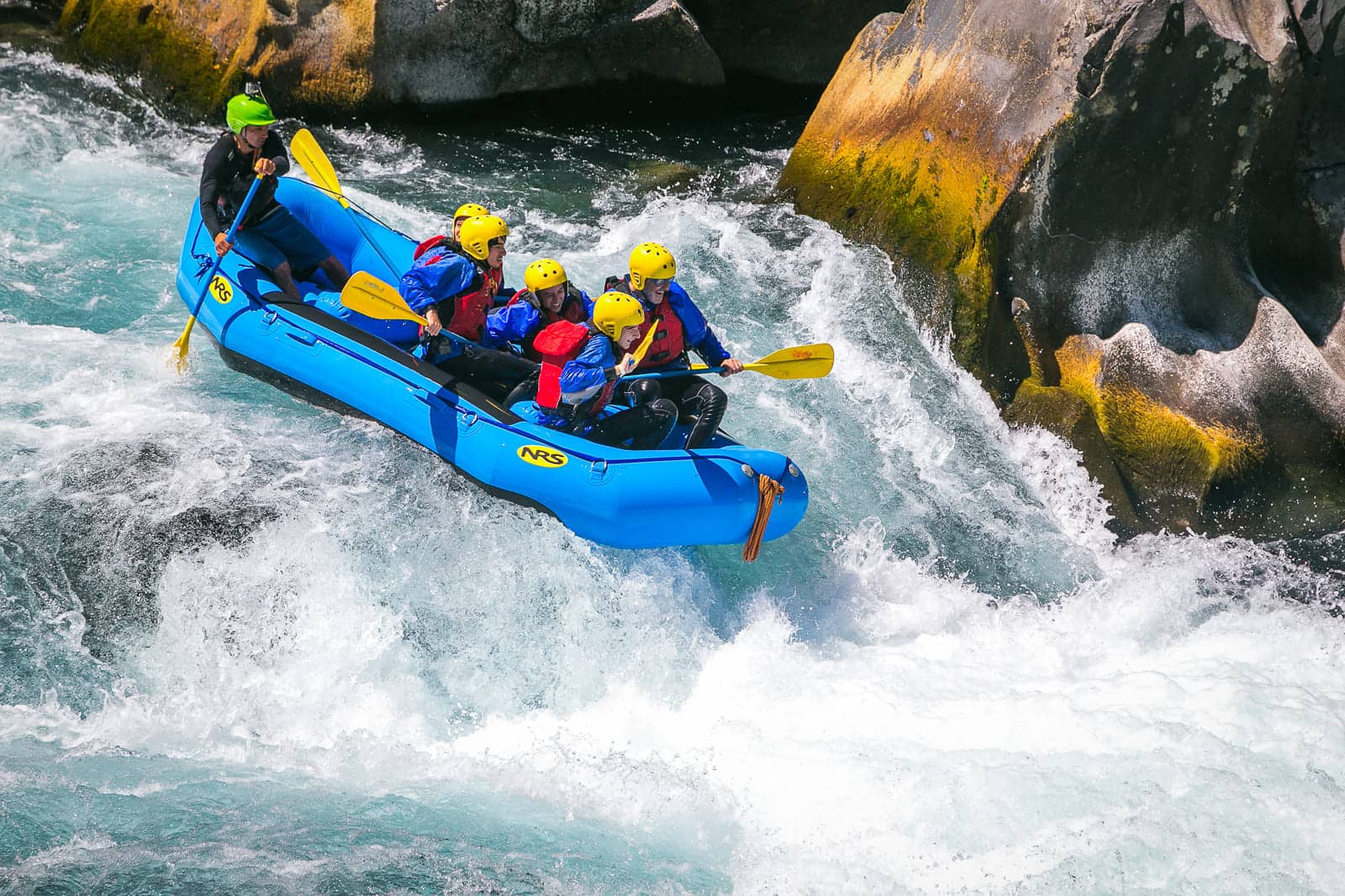 People wearing life vest and yellow helmets are rafting down a rapid river in a blue raft.