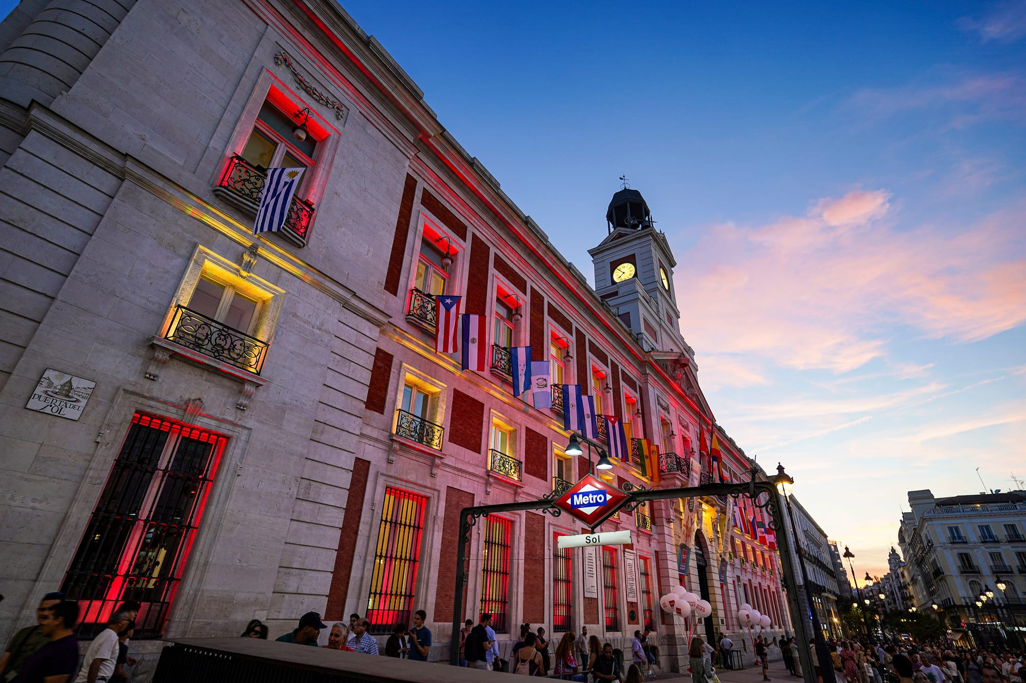 The historical building of Real Casa de Correos at sunset is lit up with red lights in the windows and people are standing near by.