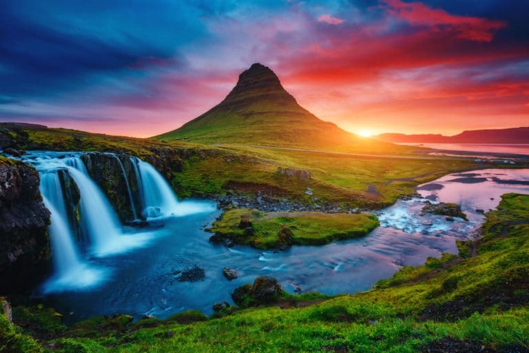 Scenic view of Kirkjufell mountain on the Snæfellsnes Peninsula in Iceland, with a vivid sunset sky in hues of red, orange, and blue, a cascading waterfall in the foreground, and a winding river flowing through lush green landscape.
