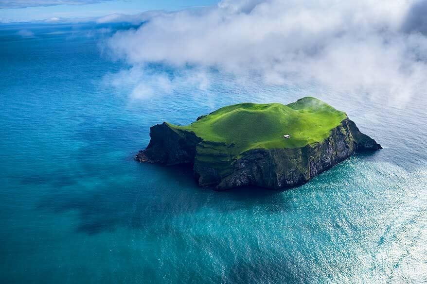 Aerial view of a small, lush green volcanic island surrounded by vibrant blue ocean waters, with dramatic cliffs and a single white house near the center. Low clouds drift above the sea, creating a dreamy, remote atmosphere.