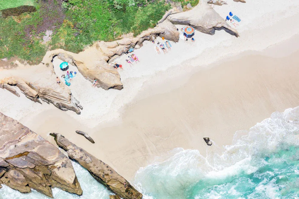  Aerial view of Windansea Beach coves in La Jolla, California, with sunbathers relaxing under umbrellas between rocky formations on soft sand next to turquoise waves.
