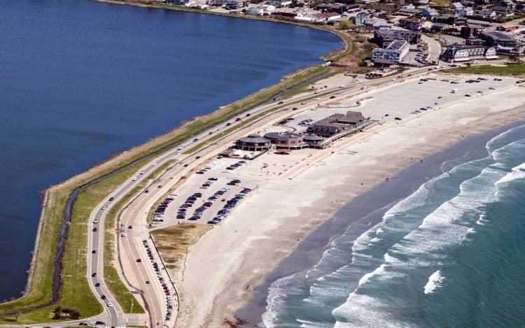 A beach with a road that runs behind it and waves washing a shore.