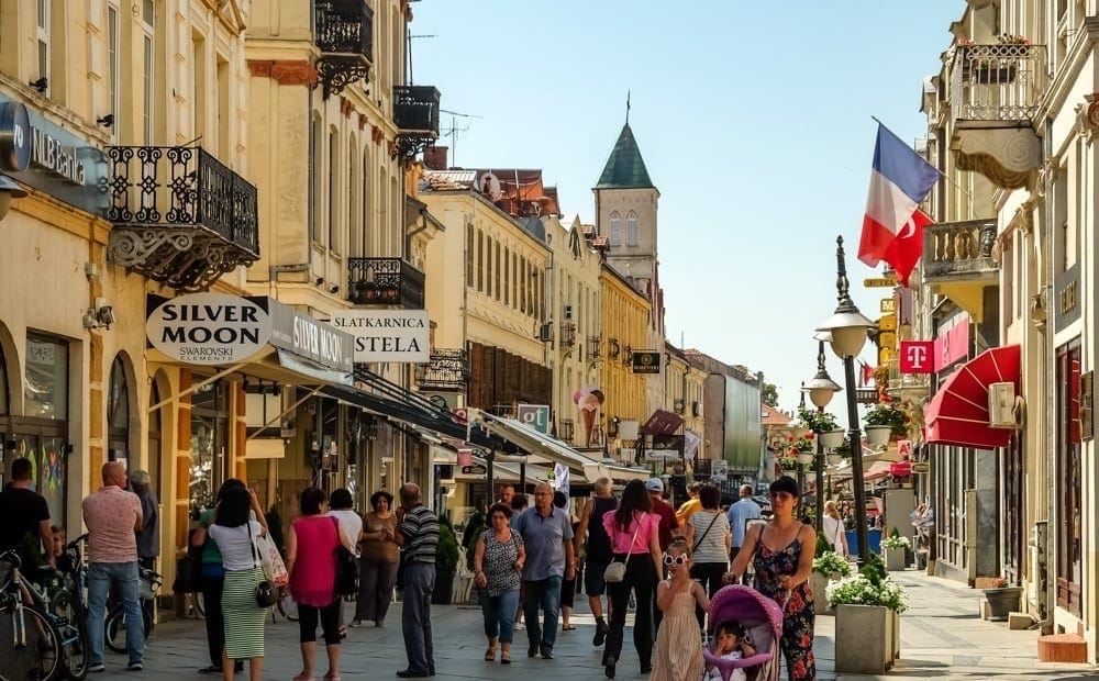 Evening street scene in Bitola, North Macedonia, featuring historic European-style buildings, shops with illuminated signs, a cyclist in the foreground, and a tower with a spire under a twilight sky with birds flying overhead.