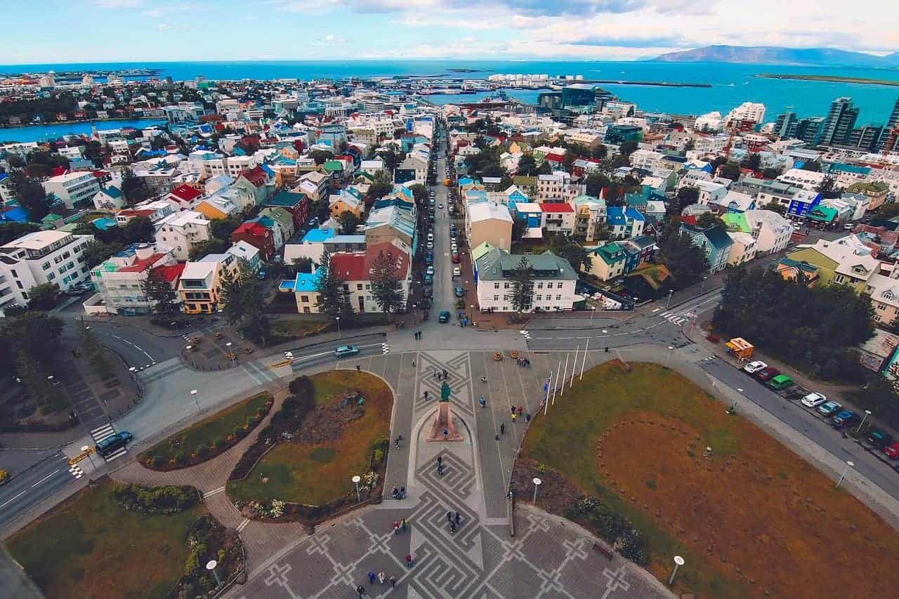 Aerial view of downtown Reykjavik, Iceland, showcasing colorful rooftops, tree-lined streets, and coastal harbor views, with a geometric plaza and statue in the foreground leading toward the city center and blue North Atlantic Ocean in the background.