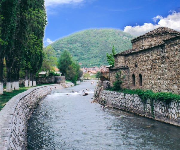 Scenic view of a stone-paved riverbank with a clear stream flowing through a historic town, featuring an old stone building on the right and lush green trees on the left, with a forested hill and blue sky with clouds in the background.