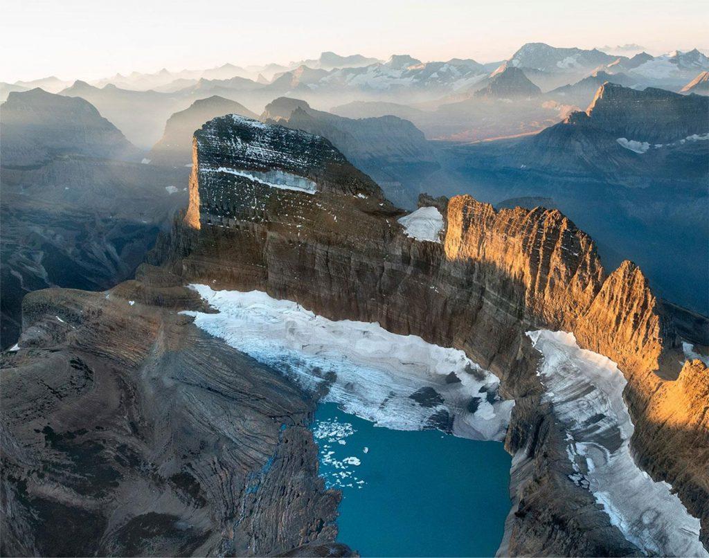 Aerial view of Grinnell Glacier and turquoise glacial lake surrounded by rugged peaks in Glacier National Park, Montana, at sunrise.
