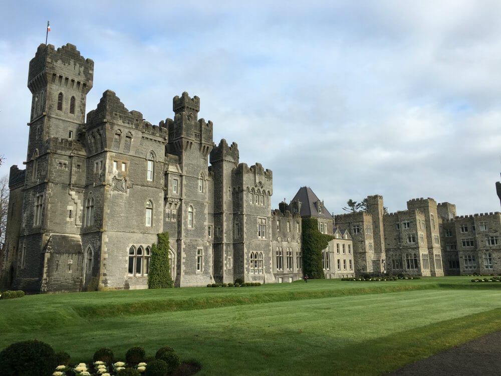 Majestic Ashford Castle in Ireland featuring tall stone towers and ivy-covered walls, set against a cloudy sky with manicured green lawns in the foreground.